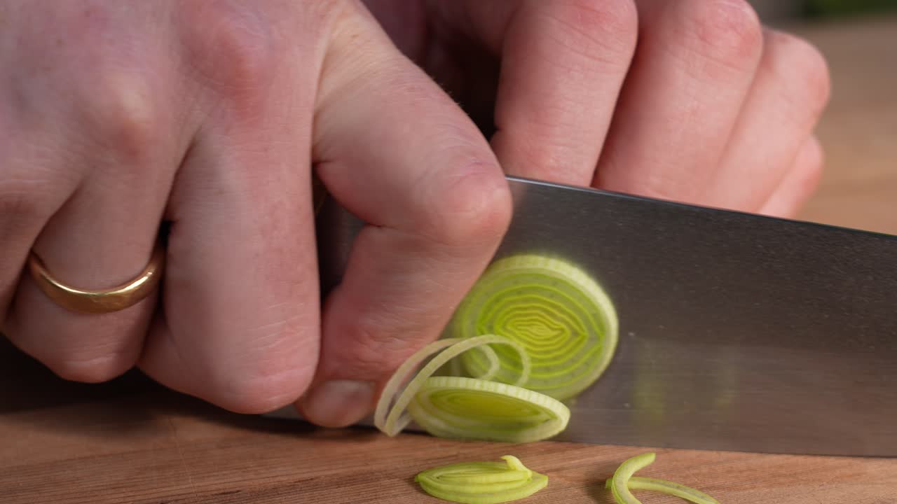 A close-up shows fresh leek being cut into thin rings on a kitchen board. Smooth knife motion reveals bright green layers and clean vegetable texture during careful preparation