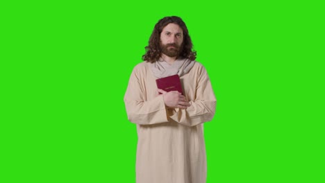 studio shot of man wearing robes with long hair and beard representing figure of jesus christ holding bible on green screen