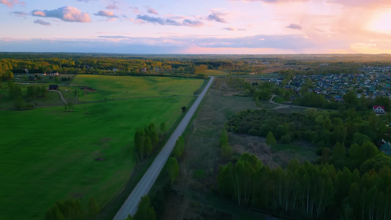 Drone view of a long rural road slicing between lush fields and forest edge under a pastel sunset near Malutki. Malutki, Latvia (Malutki, Latvija)
