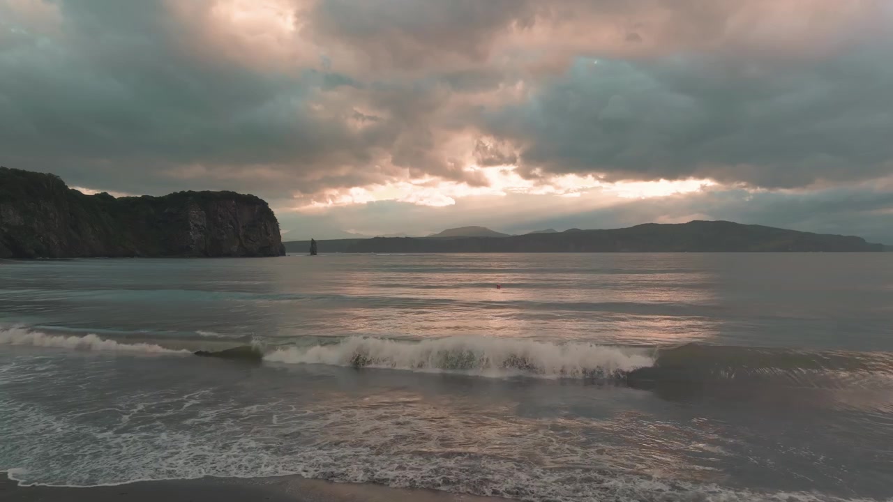 Filmagem de paisagem marinha cênica com ondas suaves rolando em uma praia de areia sob nuvens de tempestade sombrias e luz quente do pôr do sol, criando uma atmosfera costeira tranquila