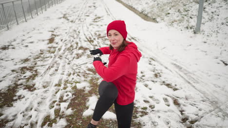 adult performing energetic winter workout outdoors, lifting leg with hand for balance on snowy ground near stairway, wearing red hoodie, surrounded by snowy hill, pathway, and foggy atmosphere