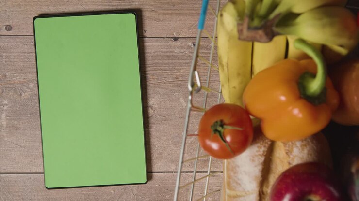 Overhead Studio Shot Of Person Using Green Screen Digital Tablet Next To Basic Food Items In Supermarket Wire Shopping Basket
