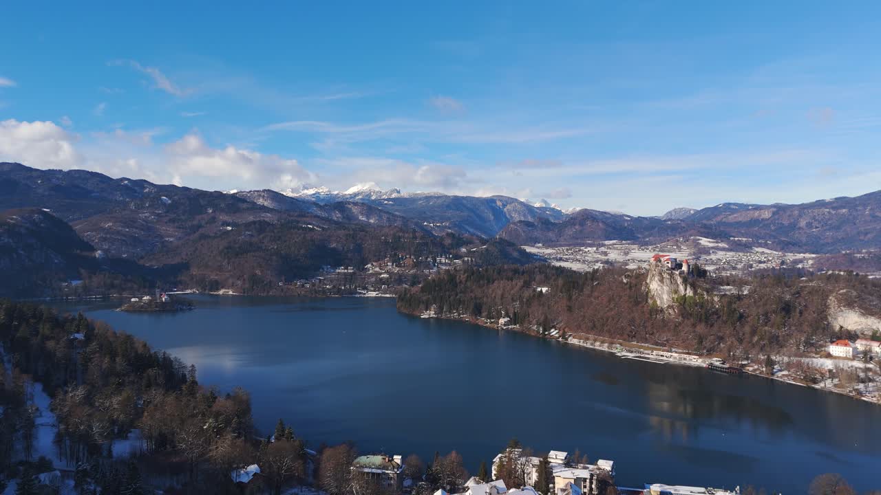 Il Lago di Bled e il Castello di Bled appaiono sotto montagne innevate in Slovenia, suggestiva ripresa aerea invernale con drone in allontanamento.