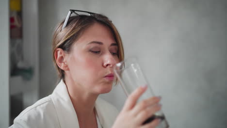 elegant woman with eyeglasses on head and hair tied back drinks coffee from transparent glass during short break in office setting, enjoying refreshment