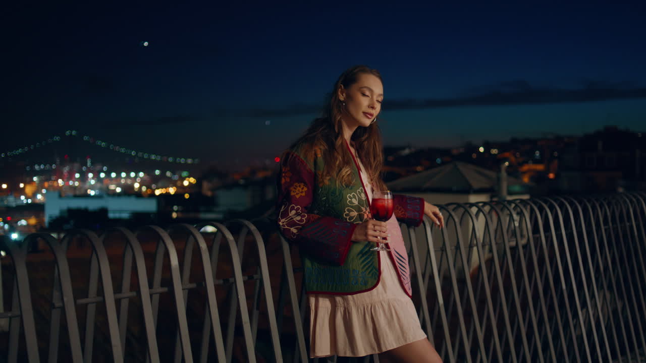 расслабленная девушка пьет коктейль, стоя у городского забора ночью. женщина наслаждается городом