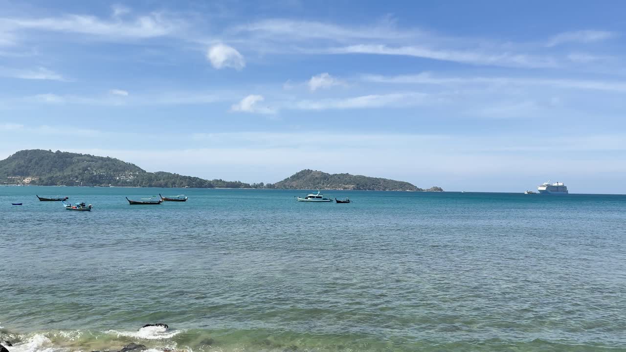 Daytime camera pan across clear sea, fishing boats, rocky shore, distant hills, and blue sky