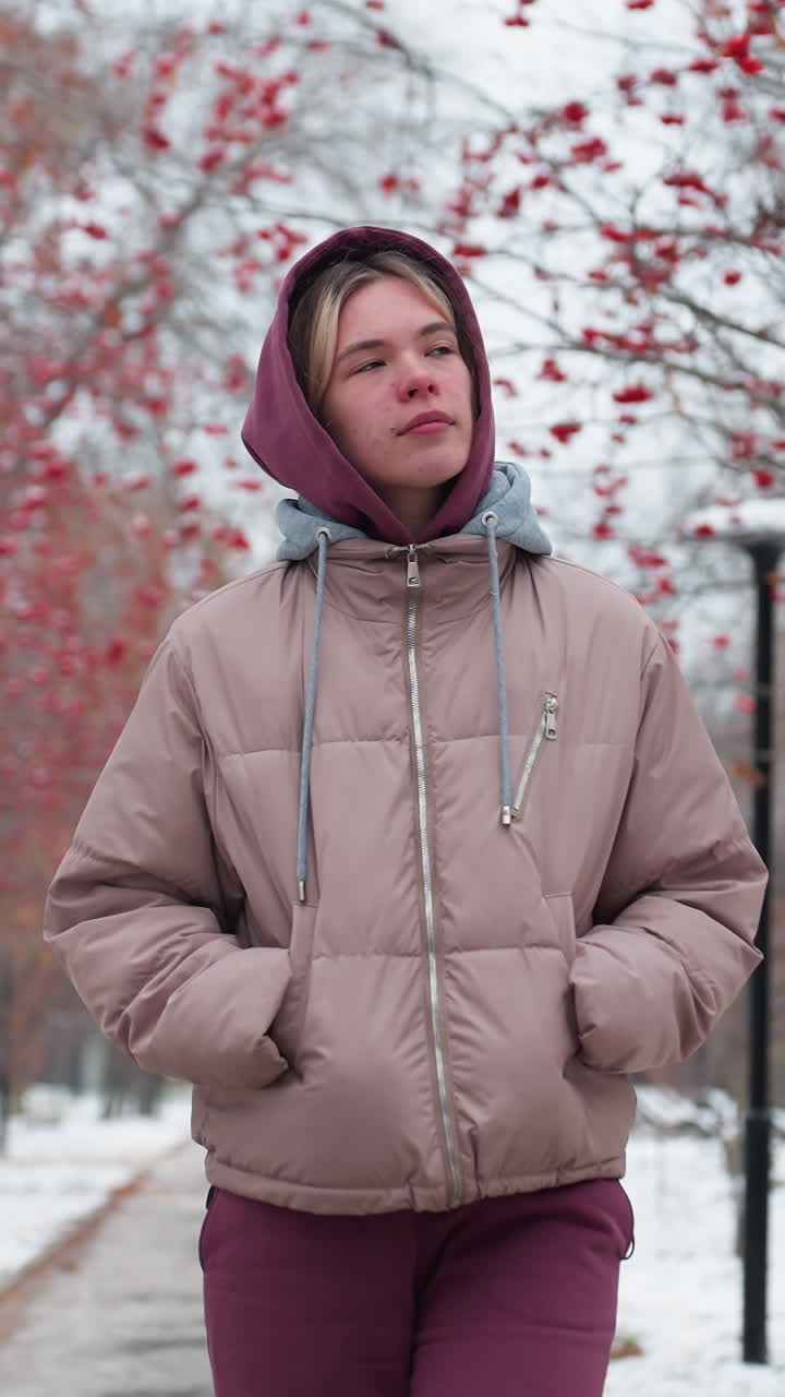 mujer joven con una chaqueta con capucha cálida camina por un camino nevado con las manos en los bolsillos, mirando pensativo, árboles de bayas rojas en línea pacífico parque de invierno