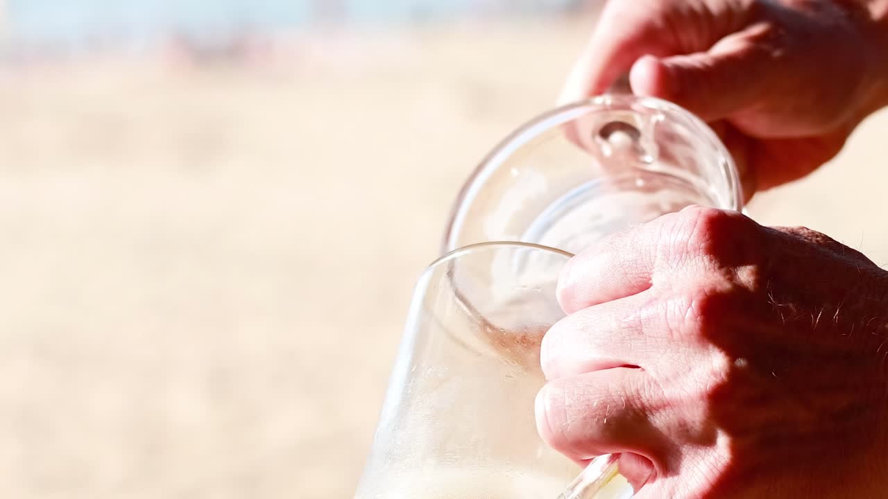 Close-up of hands pouring beer from a pitcher into a glass on a sunny beach.