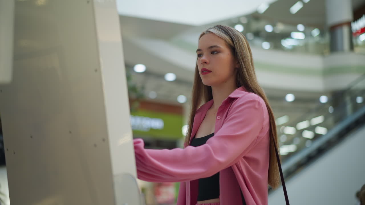 Lady in pink dress with black hand bag intensely focused on using a screen in a well-lit mall, the background features blurred lights and escalators, she appears deep in concentration