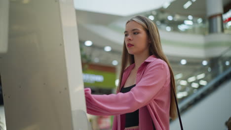 lady in pink dress with black hand bag intensely focused on using a screen in a well-lit mall, the background features blurred lights and escalators, she appears deep in concentration