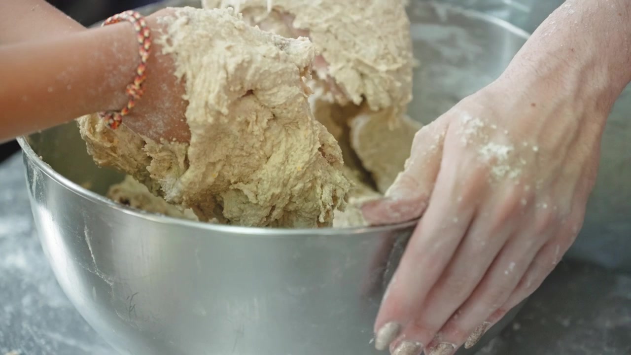 Close view of a woman's hands with long manicured nails and a beaded bracelet working a soft dough in a stainless steel bowl on a floured counter.
