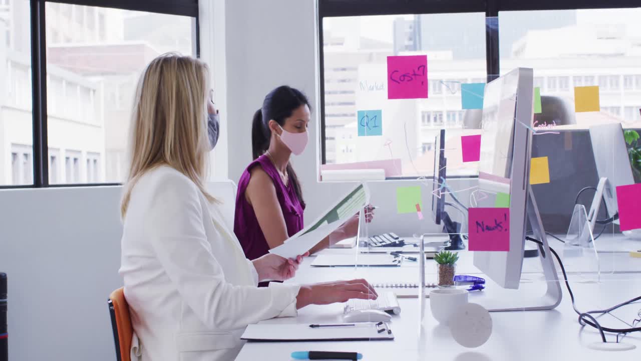 mujeres de negocios diversas que usan máscaras faciales usando computadoras pasando papeleo en la oficina