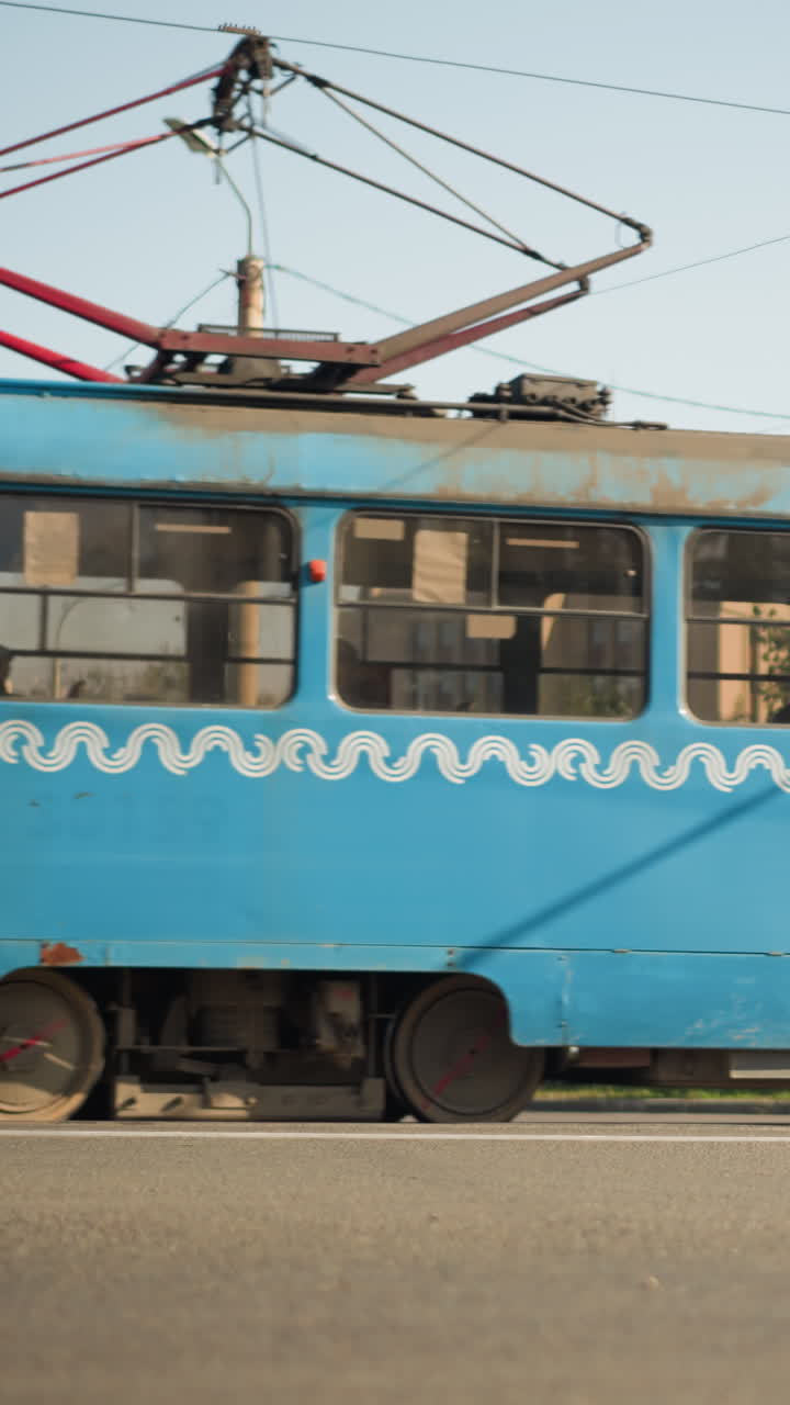 전차 한 대가 머리 위 전선과 가로등이 얽혀 있는 조용한 도시의 거리를 지나가고, 아스팔트 도로, 푸른 나무, 멀리 보이는 돔형 건물로 둘러싸여 있으며, 정오의 햇빛이 부드럽고 향수를 불러일으키는 빈티지한 광채를 드리우고 있어요.