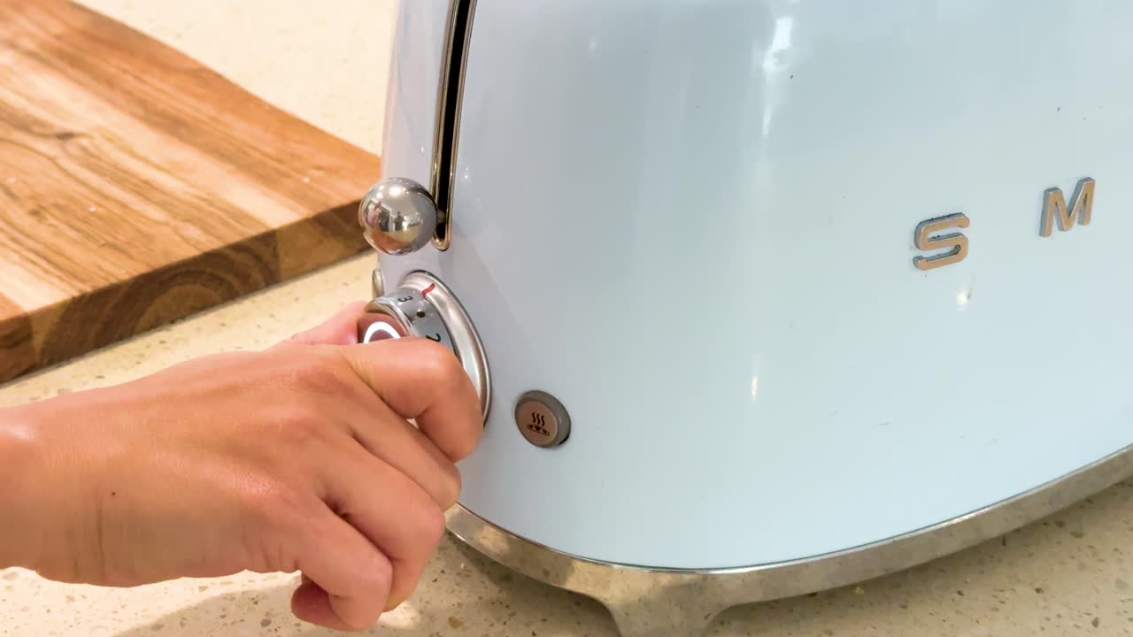 A hand rotates the dial on a retro-style toaster in a bright kitchen, adjusting settings. Natural lighting, close-up shot, steady camera, neutral mood