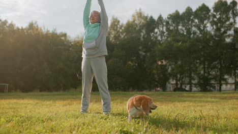 tamer in mint and grey tracksuit standing on grassy park raising hands upward eyes closed performing morning stretching exercise beside beagle dog under soft sunlight