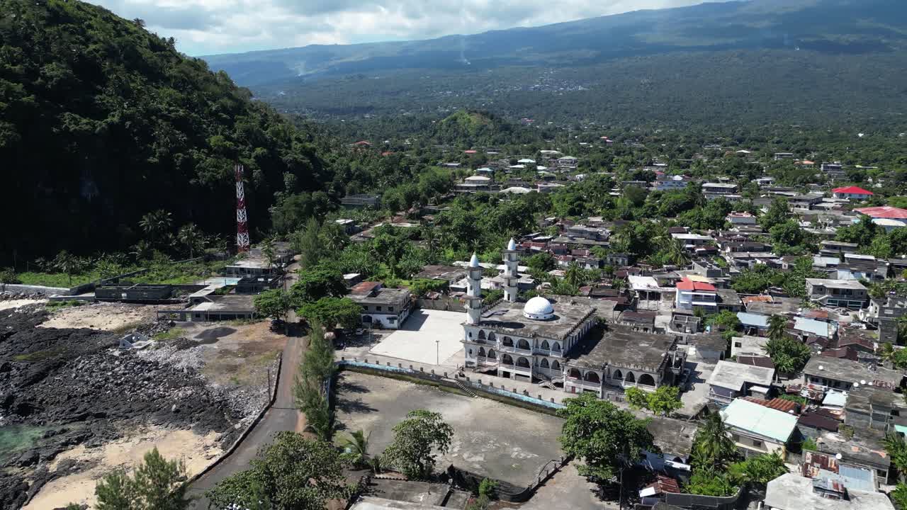 Aerial shot of coastal town with a mosque and rocky shoreline with lush green trees.