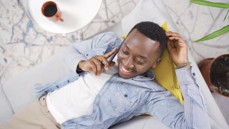 happy and cute african young man lying on sofa at home and talking on the phone.