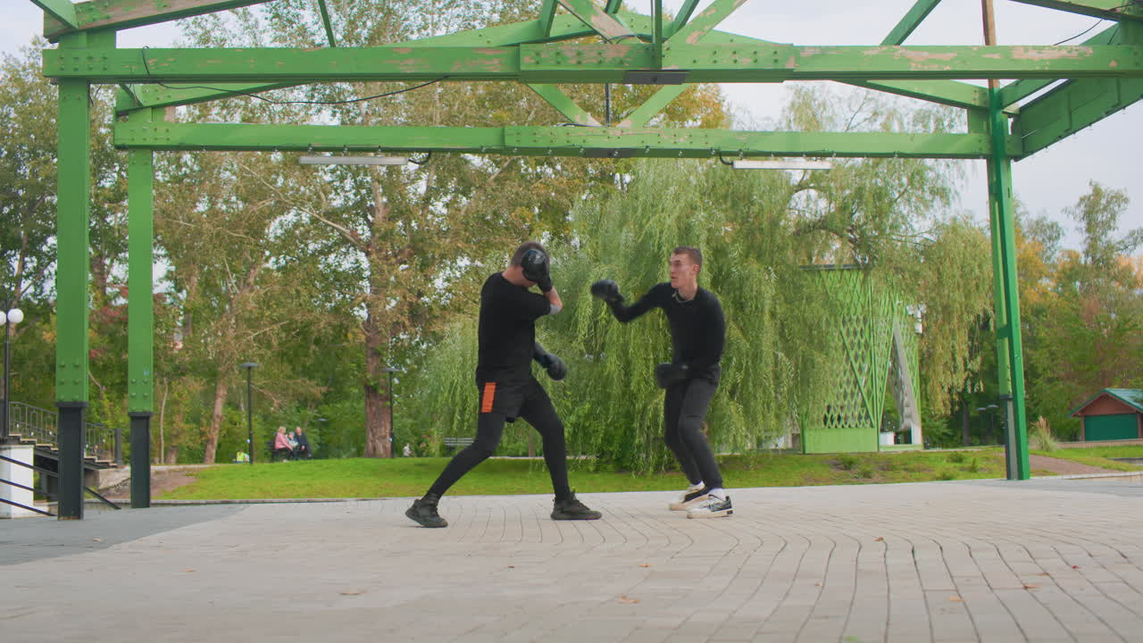 due uomini bianchi che si allenano sotto un pergolato in un parco, allenatore e atleta si esercitano con esercizi di boxe con jab veloci e un gioco di gambe preciso, struttura in metallo verde e salici sullo sfondo, superficie pavimentata e autunno