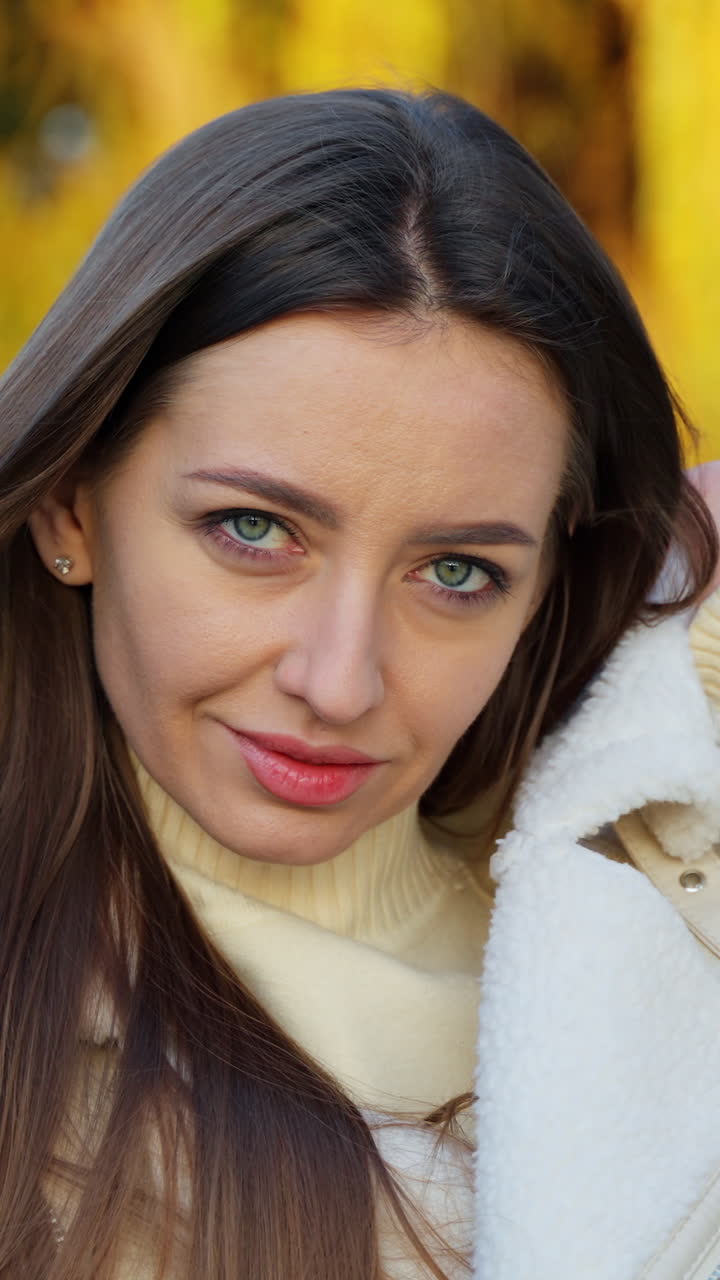 Gorgeous brunette with beautiful green eyes in the rays of bright autumn sun. Woman stands outdoors at the backdrop of yellow trees in blur. Vertical video