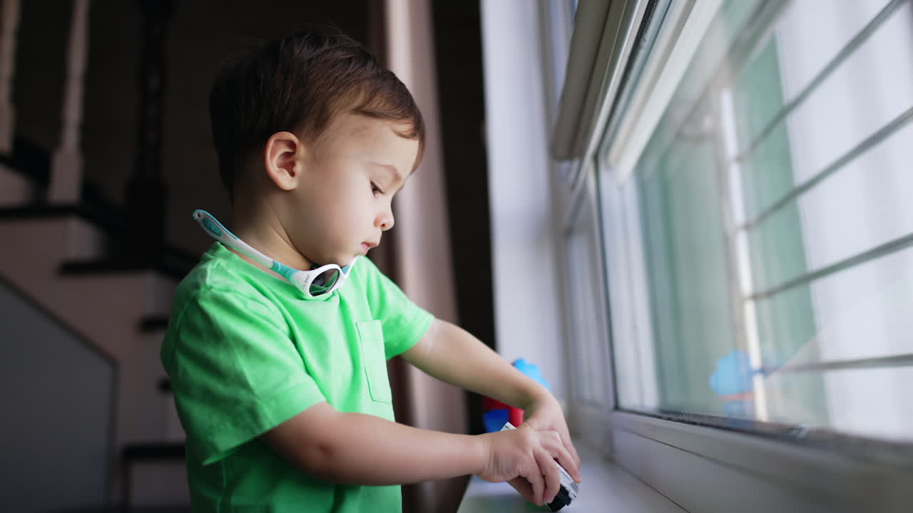 Cute toddler with plump cheeks stands at the window holding a toy. Lovely baby boy playing with a car. Blurred backdrop.