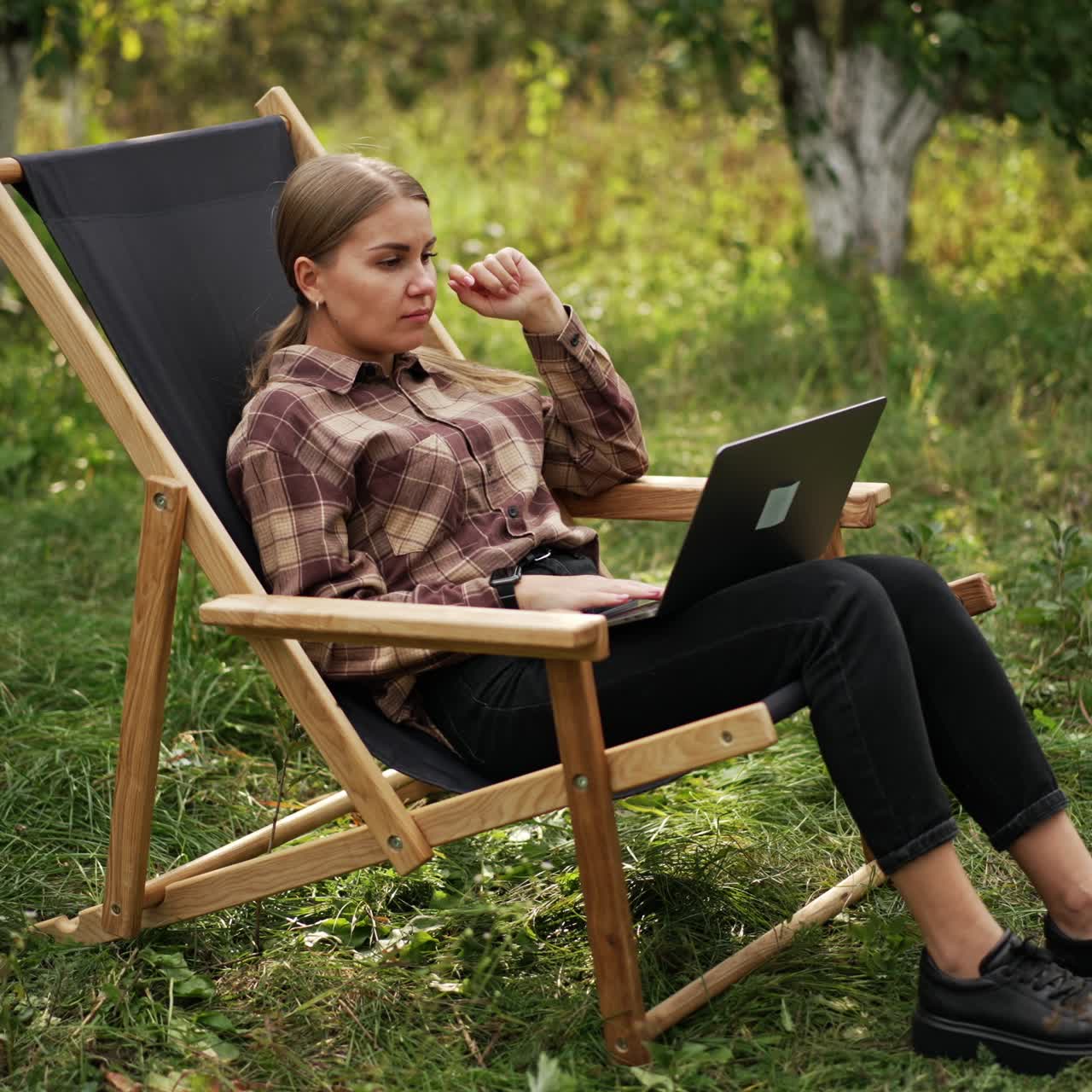 Giovane freelance che lavora al computer portatile in giardino. La ragazza si ferma un attimo a riflettere su qualcosa e poi continua a lavorare felicemente.