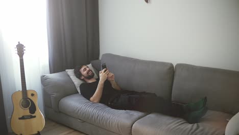 Young handsome guy relaxing on sofa using mobile, enjoying communicating with friends