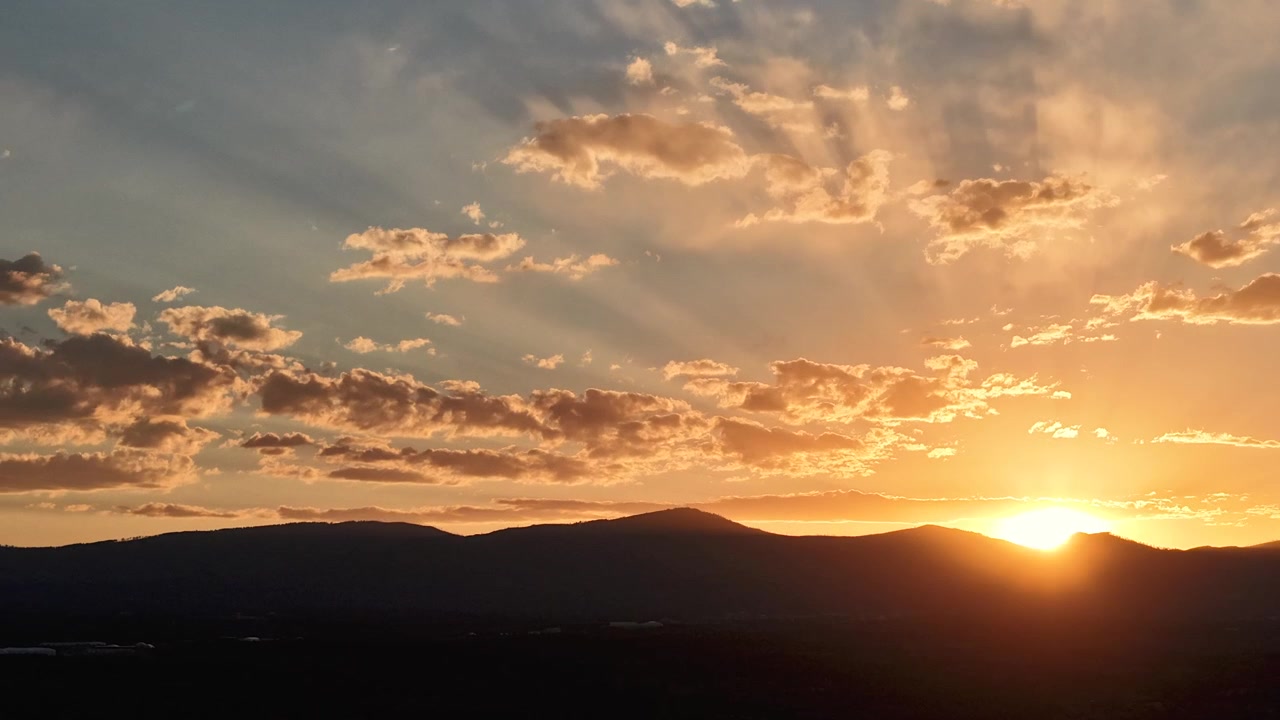 Timelapse de drone 4K UHD: ampla escala do céu captura vívidos raios de sol dourados espalhando-se por formações de nuvens dispersas. A transição atmosférica dinâmica destaca os raios de luz sobre cristas de montanhas escuras