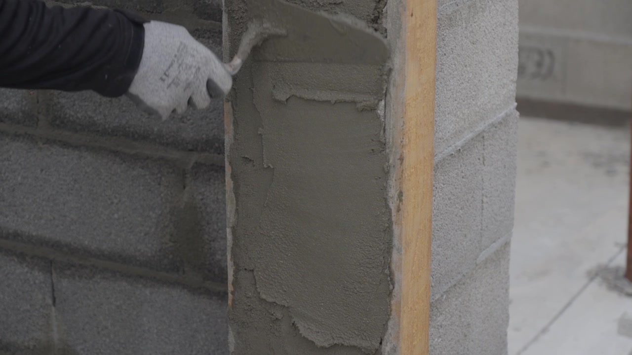 Pekerja konstruksi mengaplikasikan adukan semen segar ke dinding blok beton dengan sekop. Pekerjaan plesteran dan pasangan bata profesional di lokasi pembangunan.