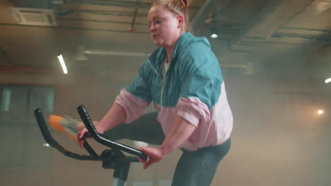 Athletic-woman-riding-on-spinning-stationary-bike-training-routine-in-haze-gym,-weight-loss-indoors