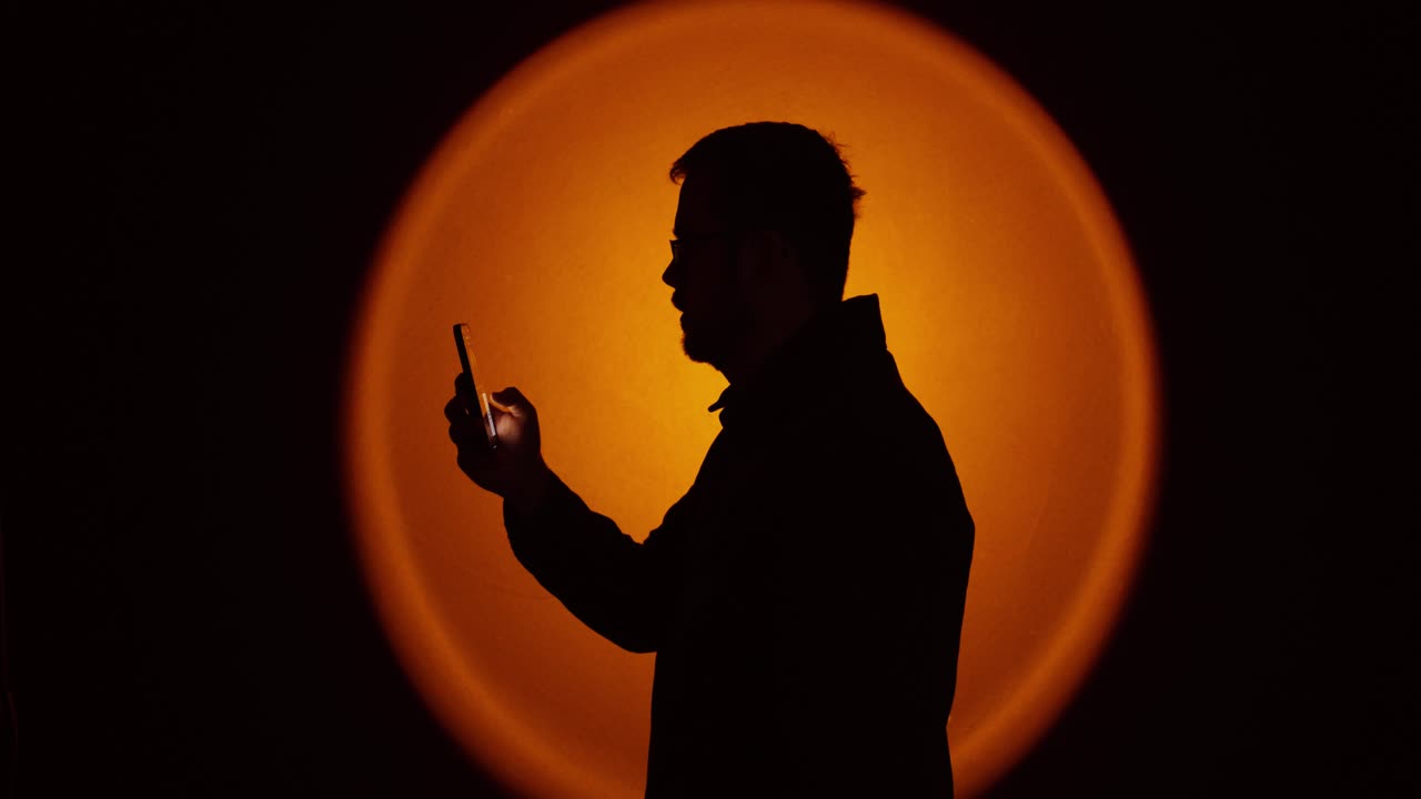 Silhouette di un uomo di profilo che guarda uno smartphone e digita, inquadrato da una calda retroilluminazione circolare in uno studio buio. Ideale per messaggistica, stile di vita digitale, privacy online, sicurezza informatica, a
