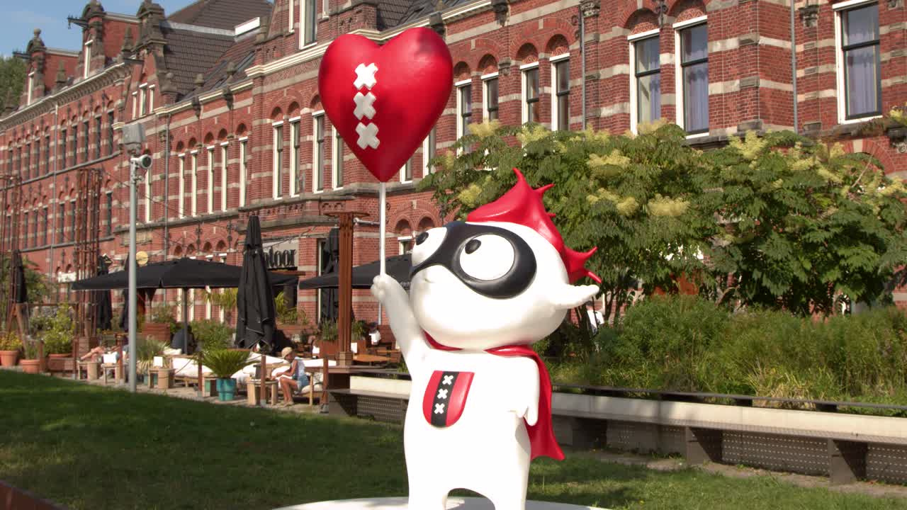 Animated mascot with heart balloon stands in sunny park near historic brick building, camera pans