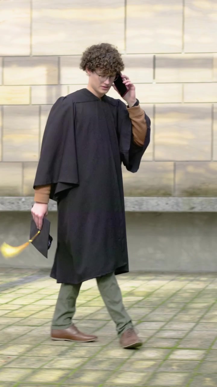 Video verticale: Uomo che cammina in toga, risponde al telefono e tiene in mano il tocco con nappa gialla in un cortile