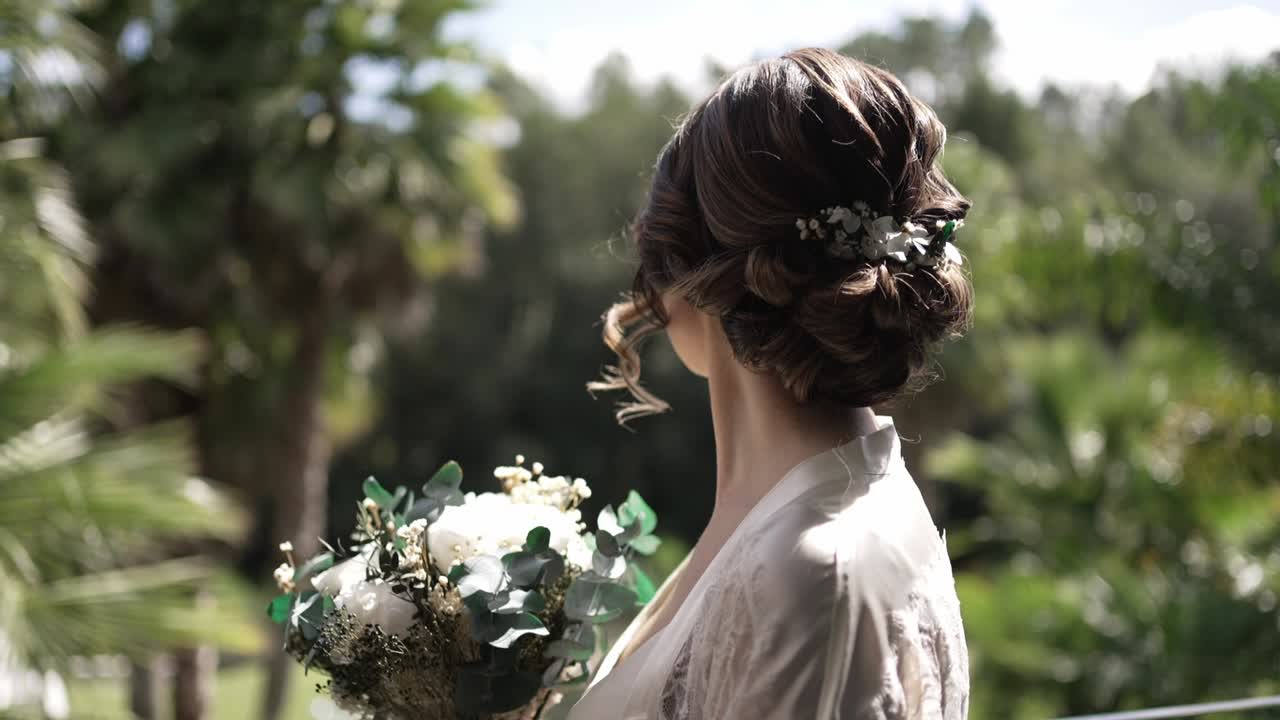 Bride with floral hairpiece holding a bouquet, looking away in a serene outdoor setting