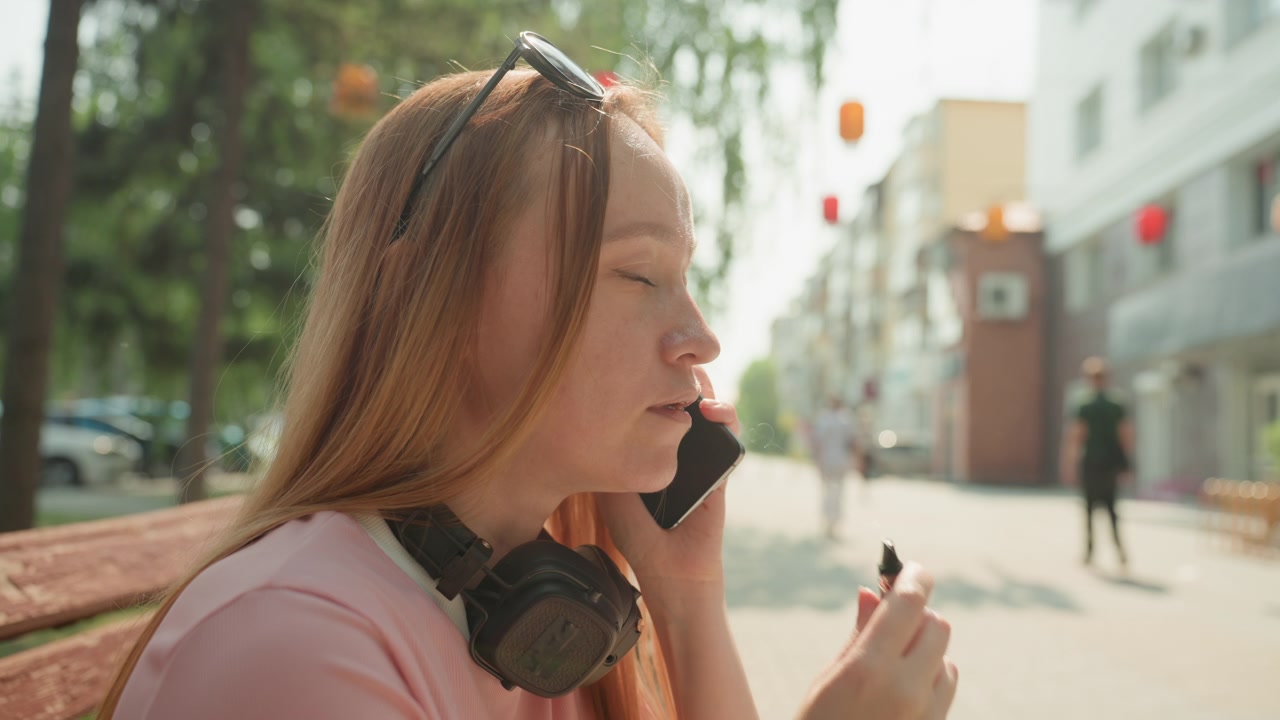 Kvinde med langt hår sidder på en bænk udendørs, holder og bruger mobiltelefon. Kvinden bærer solbriller placeret på hovedet og hovedtelefoner om halsen, engageret i samtale eller lytter.