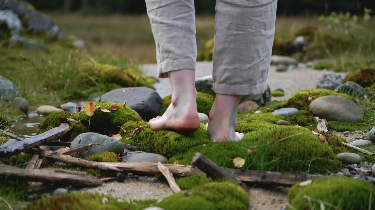 Walking barefoot adult initiating first step on narrow sandy path in cuffed trousers, passing moss