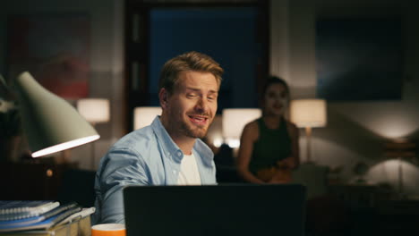 carefree woman disturbing man at dark interior. face mask wife watching laptop