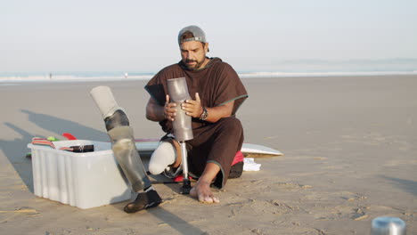 vista frontal de un surfista masculino en la playa y poniéndose una pierna protésica