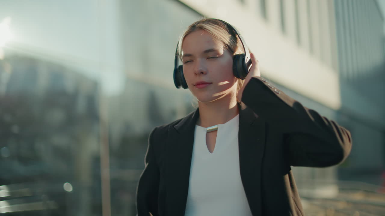 Music artist in black suit dancing while adjusting headphones and holding beverage cup, enjoying rhythm with eyes closed in urban setting surrounded by modern glass office buildings