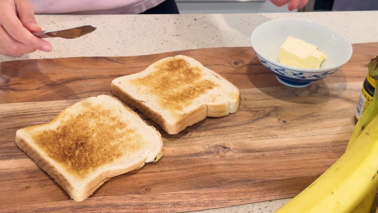 Hands spread butter on freshly toasted bread slices on a wooden board, with bananas and a butter dish visible under bright kitchen lighting