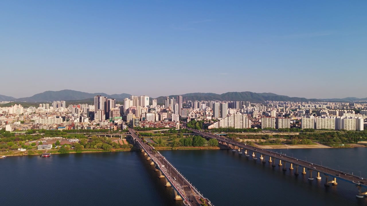 Parallasse aerea sopra i ponti Gwangjin e Cheonho che attraversano il fiume Han, con la calda luce del sole sulla densa skyline residenziale di Seul e lo sfondo montuoso