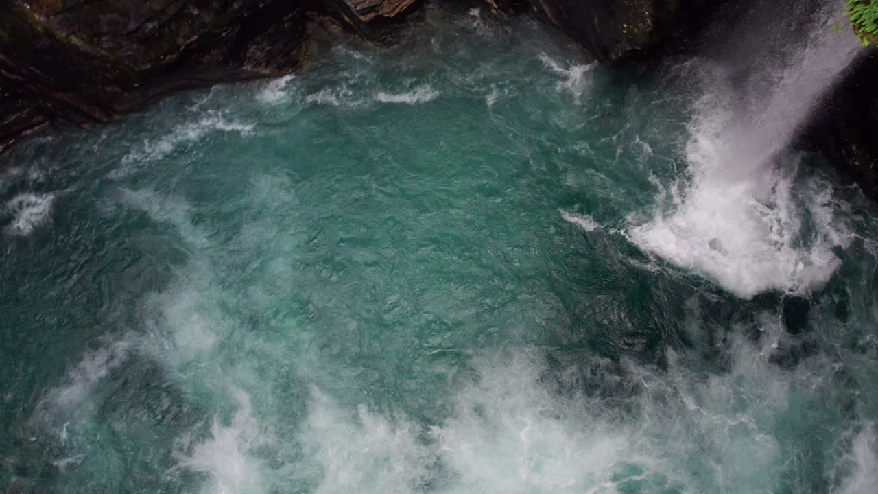 Camera descends vertically into a swirling torrent of white water in a cave, rotates and tilts upwards to reveal a waterfall pouring down through the moss covered rocks of the cave wall