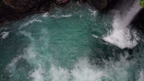 Camera-descends-vertically-into-a-swirling-torrent-of-white-water-in-a-cave,-rotates-and-tilts-upwards-to-reveal-a-waterfall-pouring-down-through-the-moss-covered-rocks-of-the-cave-wall