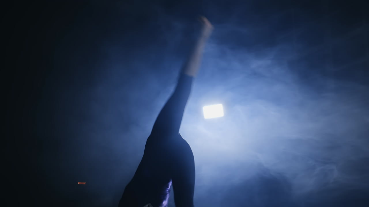 Close-up of a Talented girl gymnast performs a flip turn in a jump on the gymnastics carpet in slow motion in a contoured light in the smoke. Steadycam