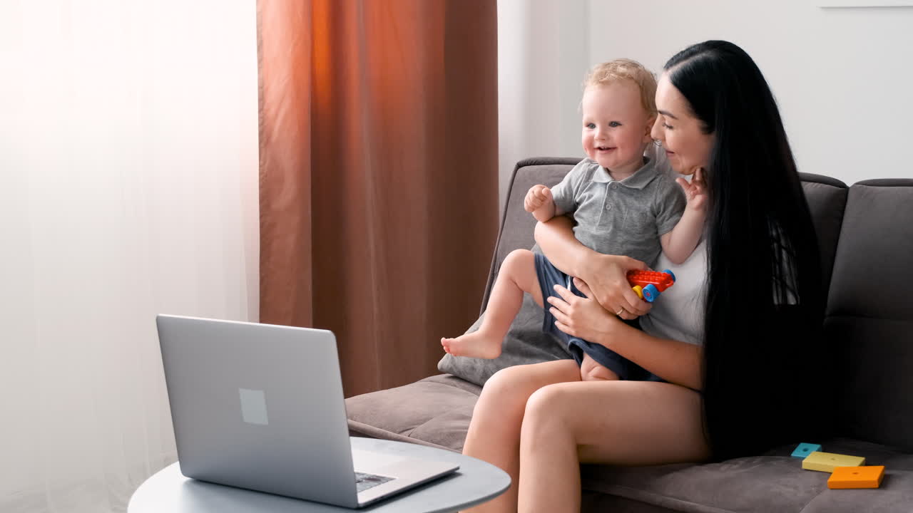 Beautiful Mom Sitting On Sofa In Living Room Holding Her Adorable Little Boy While Having A Video Call On Moder Laptop At Home