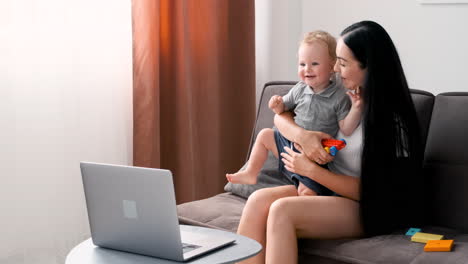 beautiful mom sitting on sofa in living room holding her adorable little boy while having a video call on moder laptop at home