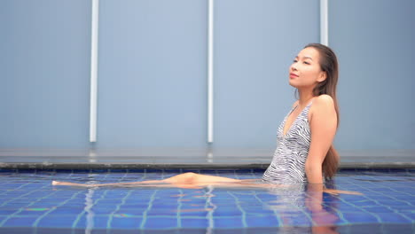 Thai-Woman-in-Zebra-Color-Monokini-Sitting-Inside-Shallow-Swimming-Pool-Water-and-Moving-Legs-Under-the-Water-Slow-Motion,-Vacation-Template