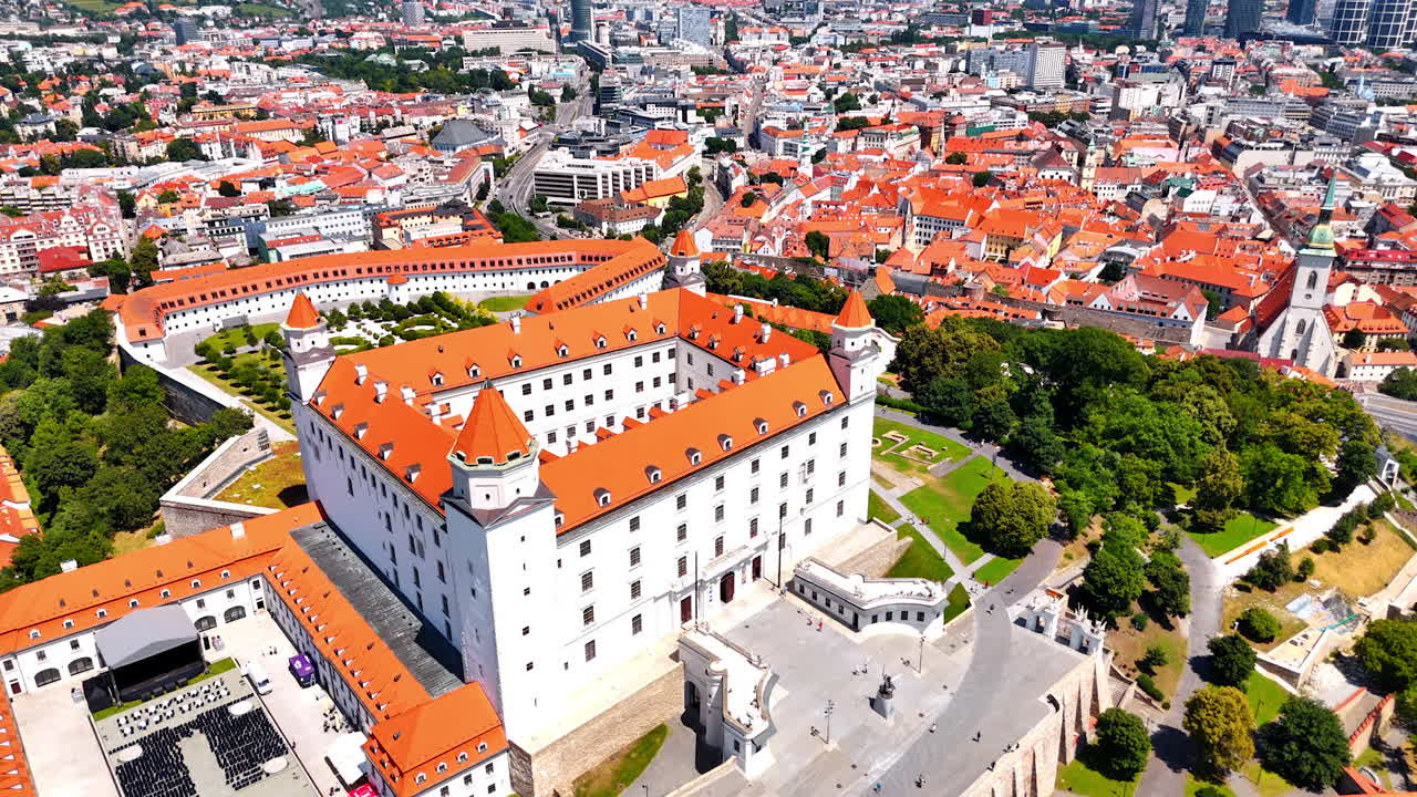 Flying over the orange roof of the Bratislava Castle. Stage and rows of chairs installed at the territory of the landmark. View of the old town at backdrop