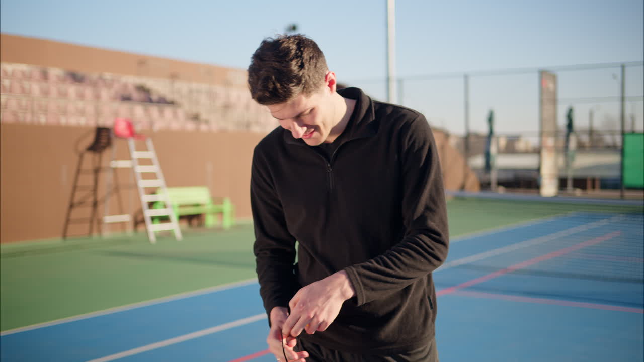 Um homem feliz e sorridente vestindo roupas pretas, segurando uma raquete de pickleball vermelha e uma bola amarela em uma quadra azul em um dia ensolarado