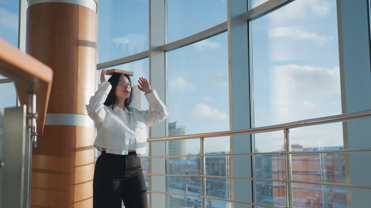 Confident woman walking through modern sunlit interior with book balanced on head, arms lifted slightly for balance, wearing white blouse and black trousers, moving gracefully beside large windows