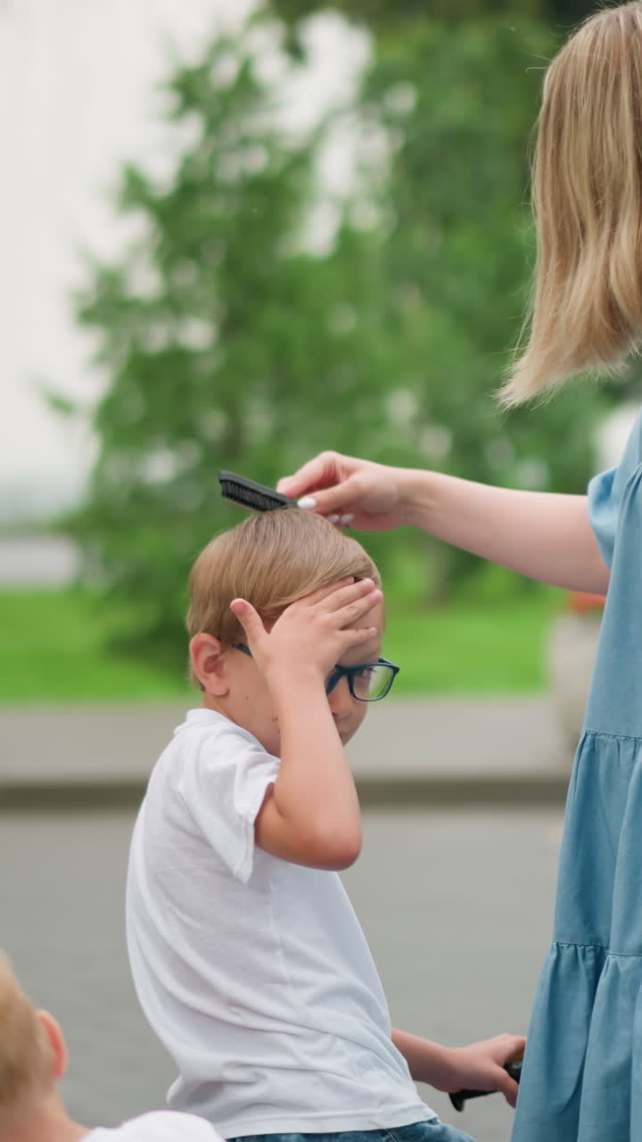 A mother carefully combs her young son s hair while he adjusts it with his hand, wearing glasses and sitting on a bicycle, his younger sibling is behind him focus on something else